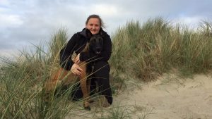 A woman hugs a dog in a sandy area with tall grass.